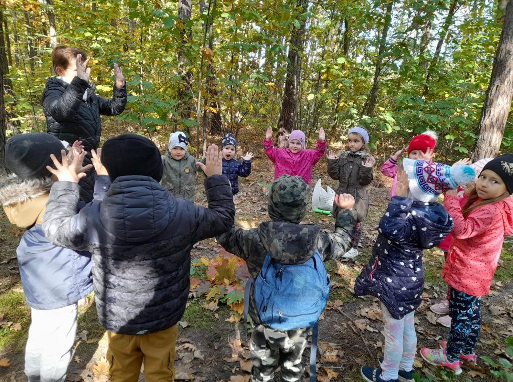 Grupa dzieci wraz z wychowawczynią stoją w kole na leśnej ścieżce. Śpiewają piosenkę podnoszą ręce do góry. Dzieci są wesołe i uśmiechnięte. W tle widać drzewa na których są kolorowe liście.