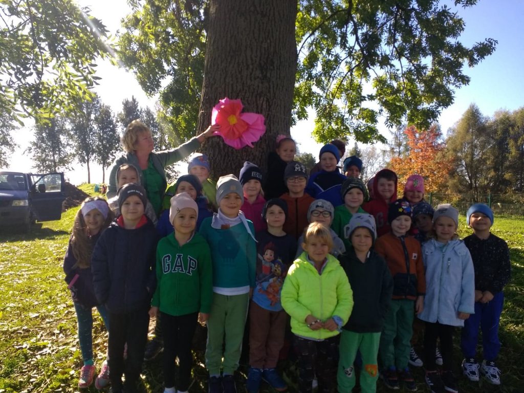 Grupa dzieci w kolorowych kurtkach stoi przed drzewem rosnącym obok boiska sportowego. Drzewo przyozdobione jest różową kokardą z czerwonym kwiatem. Jedną ręką przytrzymuje dekorację nauczycielka w zielonym ubraniu.