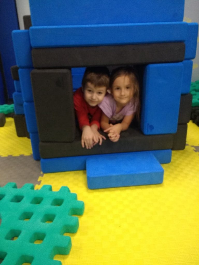 Domek zbudowany z czarno- niebieskich dużych klocków stoi na żółtej macie. W otworze domu widać dwoje dzieci: chłopca w czerwonej koszulce i dziewczynkę w różowej bluzce. Leżą opierając ciało na rękach.