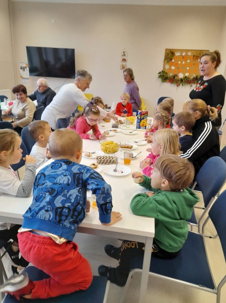 Przedszkolaki siedzą przy stolikach, dwie babcie częstują ich ciasteczkami i sokiem. W tle widać innych seniorów oraz jesienna gazetkę.