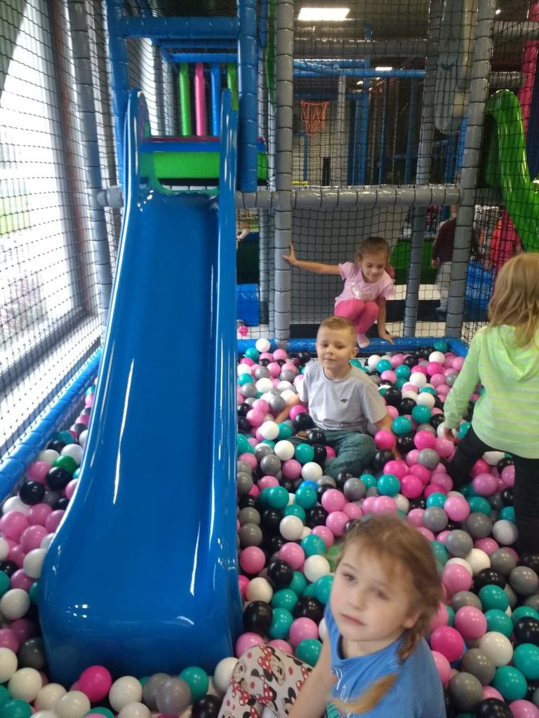 Basen z różnokolorowymi kulkami, w nim niebieska zjeżdżalnia. 3 dzieci siedzi i bawi się piłeczkami. 1 dziewczynka w seledynowej bluzie stoi z boku basenu.