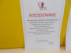 2. Podziękowanie za zbiórkę karmy - biała kartka wyeksponowana na żółtym tle.       U góry na środku znajduje się logo: napis Azyl i serce w kolorze czerwonym, w nim sylwetki kota i psa. Pod spodem czerwonym kolorem wypisane jest słowo: Podziękowanie. Pozostała treść ( w kolorze czarnym ) brzmi:
,,Dla Publicznego Przedszkola w Wietrzychowicach Przyjaciół Azylu dla psów i kotów w Tarnowie. Za gorące serca i wrażliwe na pomoc dla Braci Mniejszych umysły, albowiem prawdziwą miarą człowieka jest to, jak traktuje się kogoś, kto nie może mu się w żaden sposób odwdzięczyć. A wy ponad tą miarę wyrastacie. Z życzeniami sukcesów! Kierownik schroniska - podpis’’.
