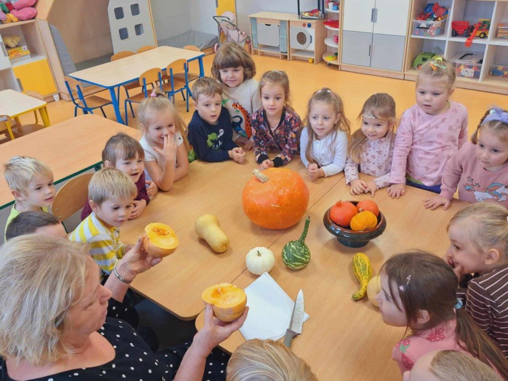 Na stole leżą dynie: duże i małe, okrągłe, podłużne, w kształcie gruszki, zielone, pomarańczowe, żółte, kremowe, białe. Dookoła stołu siedzą dzieci. Pani w czarnej sukience w białe kropki trzyma w rękach 2 połówki dyni. Pokazuje pestki w środku dyni.