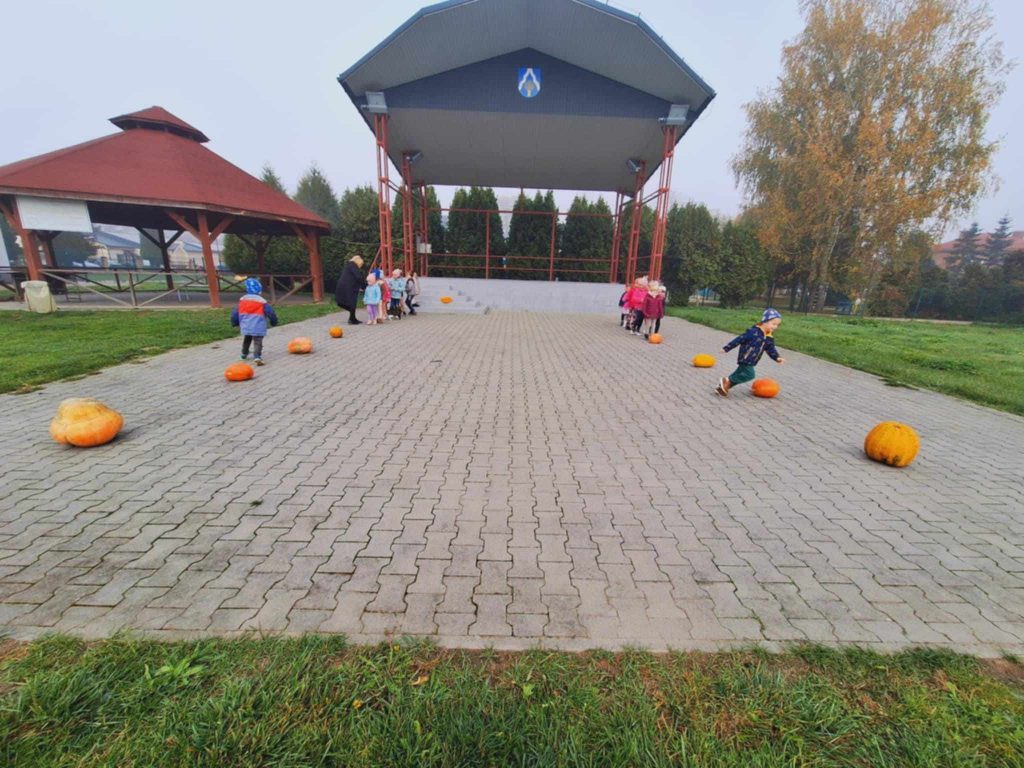 Na szarej kostce przed sceną ustawione w 2 rzędach dzieci. Obok jednego z nich stoi pani w czarnej kurtce I pochyla się nad dziećmi. Przed każdym rzędem leżą po 4 pomarańczowe dynie oddalone od siebie. Dwoje dzieci biegnie pomiędzy dyniami. Z boku drewniana altanka. Wokół zielona trawa.